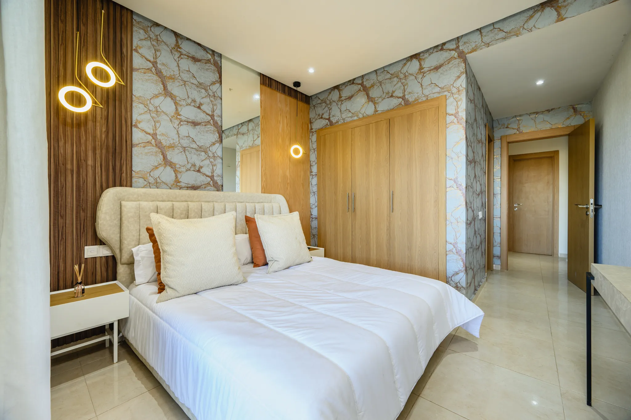 Chambre parentale élégante avec papier peint effet marbre, tête de lit capitonnée et luminaires suspendus.