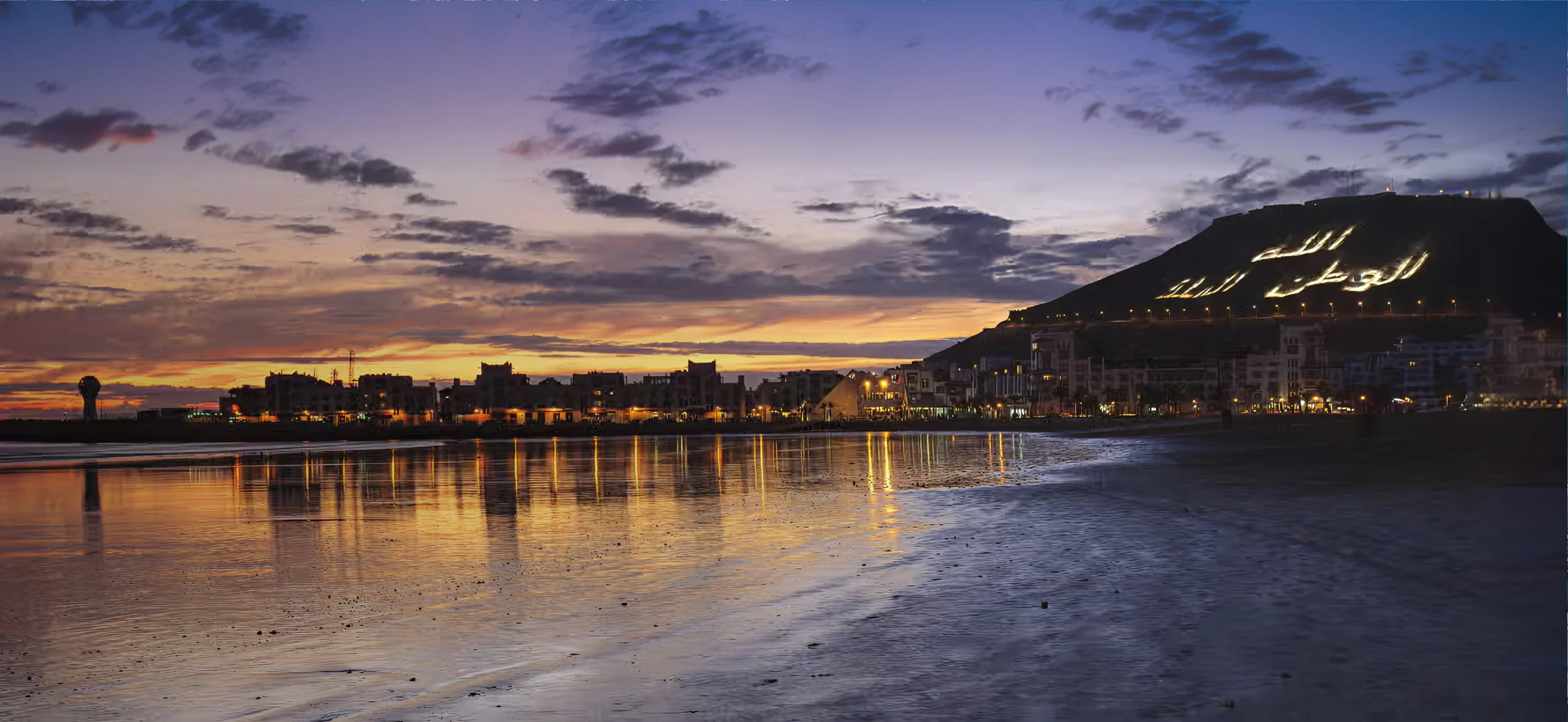 Vue panoramique de la plage d’Agadir au coucher du soleil avec la montagne illuminée et le front de mer au Maroc